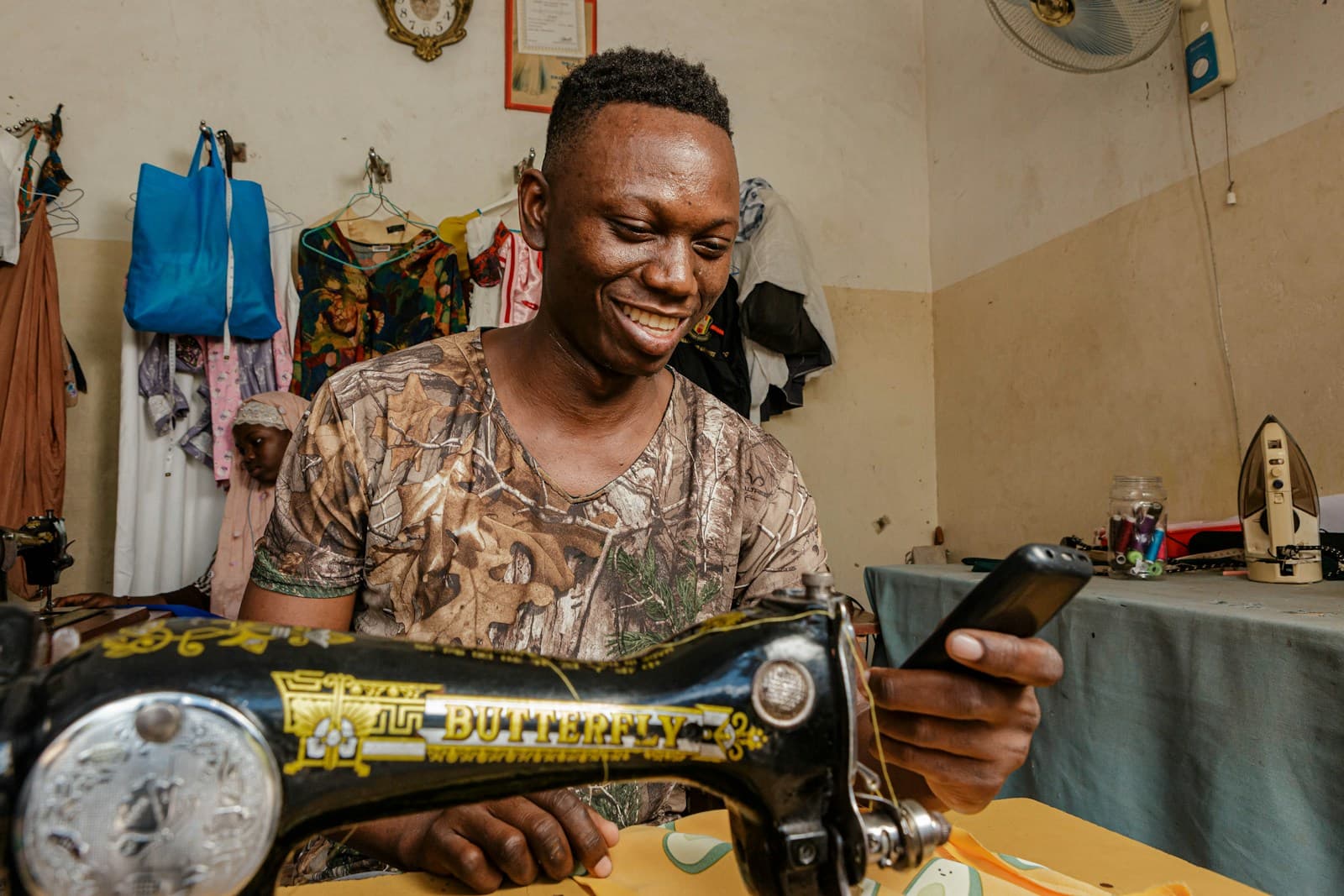 A young man smiles as he works at a sewing machine in a tailoring workshop.